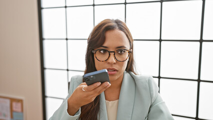 Relaxed young hispanic woman owning her business at the office, multi-tasking with a poised work-face. using her smartphone, she sends voice messages while managing work indoors.