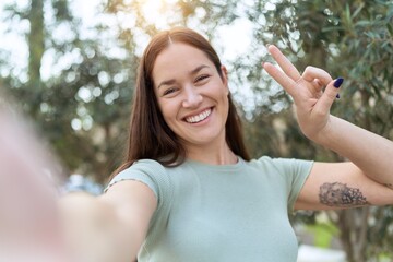 Young beautiful woman making selfie by camera doing victory gesture with fingers at park