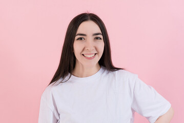Fototapeta premium Portrait of attractive Caucasian young brunette woman in casual clothes white t-shirt, smiling and looking confidently at the camera isolated on pink studio background.