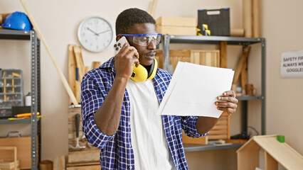 African man in a workshop talking on the phone while examining a document, wearing safety goggles...