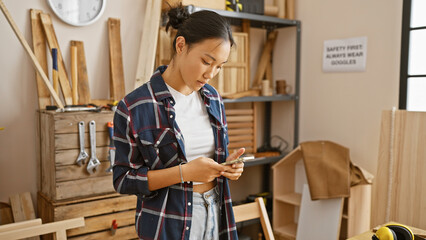 A young asian woman engaged with her mobile phone in a carpentry workshop indoor setting.