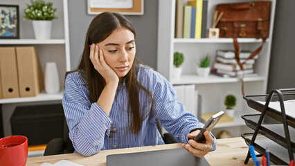 Hispanic woman with smartphone in modern office looking pensive.