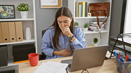 Young hispanic woman feeling stomach pain while working at her office desk, expressing discomfort.