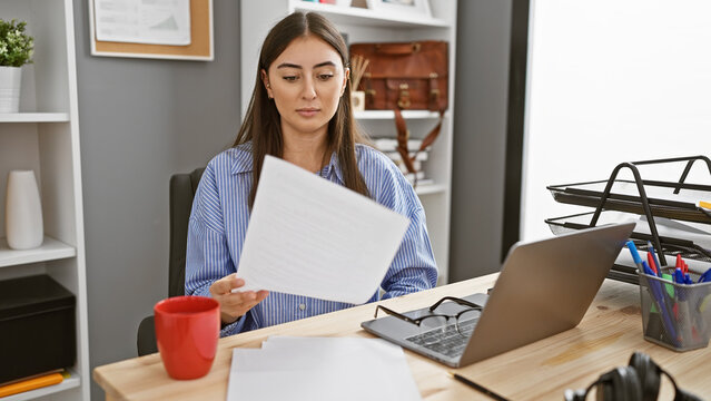 A professional young woman reviews documents at her office desk with a laptop and accessories in the background.