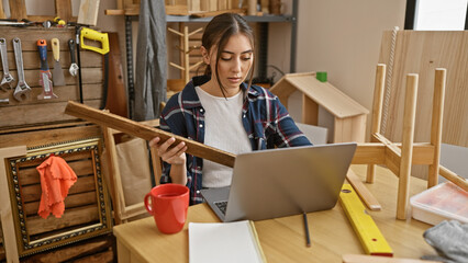 A young hispanic woman examines a piece of wood while working on a laptop in a carpentry studio filled with tools.