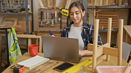 Hispanic woman uses a credit card for online payment on her laptop in a carpentry workshop
