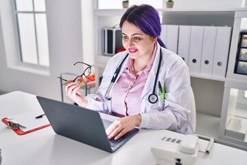 Young beautiful plus size woman doctor using laptop holding glasses at clinic
