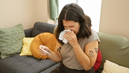 A young hispanic woman blowing her nose and checking a thermometer in a cozy living room setting