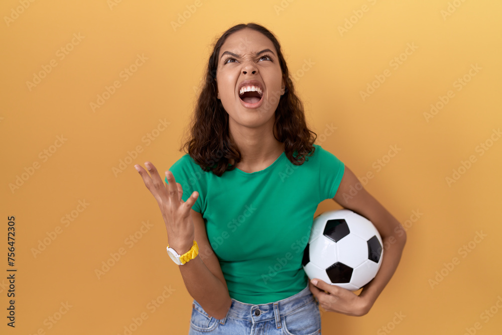 Poster young hispanic woman holding ball crazy and mad shouting and yelling with aggressive expression and 