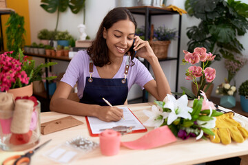 Young african american woman florist talking on smartphone writing on document at flower shop