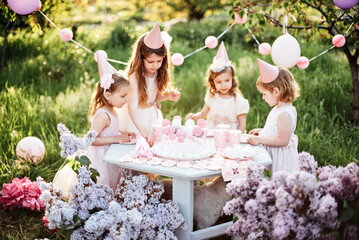 Summer outdoor kids birthday party. Group of happy Children celebrating birthday in park. Children blow candles on birthday cake. Kids party lilac pink pastel decoration and food. Presents and sweets. - 756473775