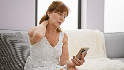Mature woman with red hair experiencing neck pain while looking at smartphone in cozy living room.