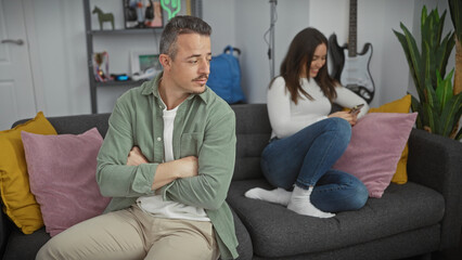 A displeased man and a distracted woman with a phone sitting on a couch in a modern living room.