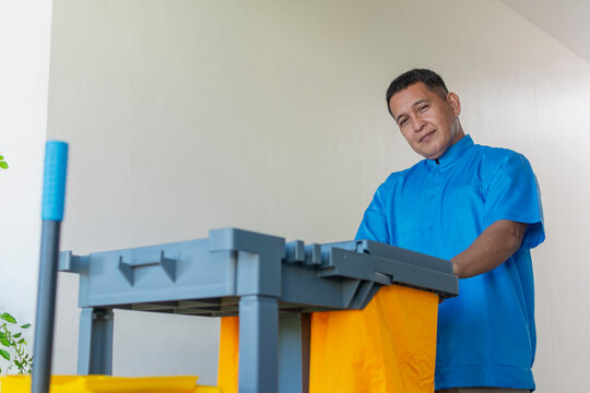 A dedicated middle-aged male janitor in a blue uniform pushes a cleaning cart along a bright hallway.