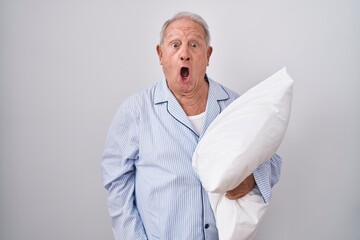 Senior man with grey hair wearing pijama hugging pillow in shock face, looking skeptical and...