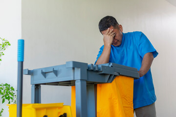 Middle aged janitor showing signs of stress and fatigue while pushing a cleaning cart down a...