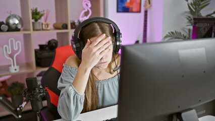 A stressed young woman in a gaming room, wearing headphones, with a microphone and a computer,...