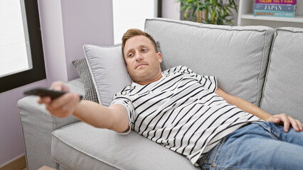 Bored young caucasian man, lounging on the sofa, hopelessly channel surfing at home