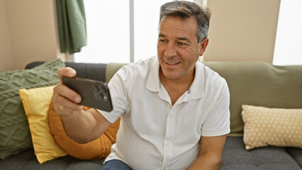 Smiling middle-aged man using smartphone in cozy living room, portraying connection and relaxation.