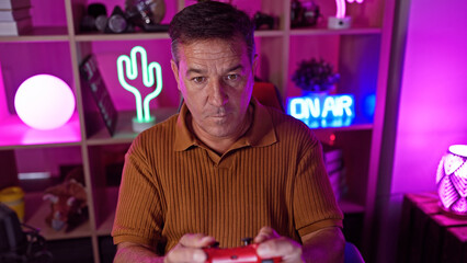 A middle-aged man engages in videogaming, holding a controller in a cozy, neon-lit room at night,...