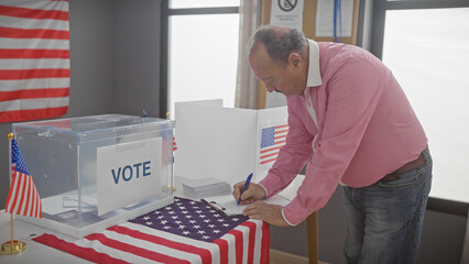 A mature man in a pink shirt writes at a polling station with us flags and a ballot box indicating a voting scene. - obrazy, fototapety, plakaty