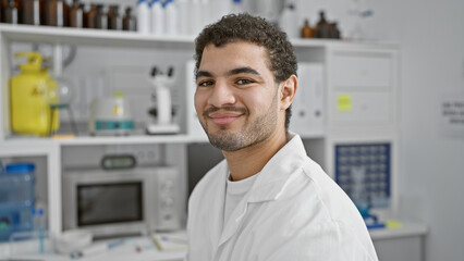 Naklejka na ściany i meble A confident man with a beard in a lab coat stands in a well-equipped laboratory, smiling at the camera.