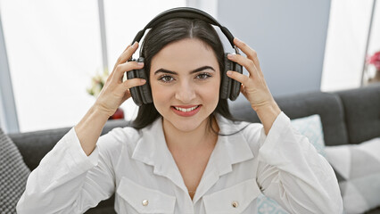 A smiling young hispanic woman enjoys music on headphones in a cozy living room