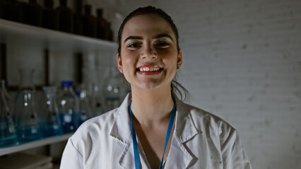 A cheerful hispanic woman in a lab coat stands in a laboratory with glassware shelves, exemplifying...