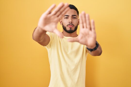 Young hispanic man standing over yellow background doing frame using hands palms and fingers, camera perspective