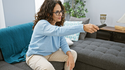 A mature hispanic woman with curly hair uses a remote control in a cozy living room setting.