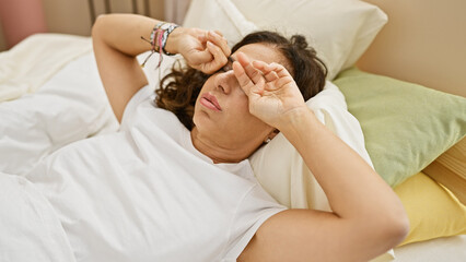 A middle-aged hispanic woman with curly hair lies in bed indoors, looking tired or unwell, rubbing...