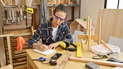 A mature hispanic woman meticulously takes notes in a well-equipped carpentry workshop, surrounded...