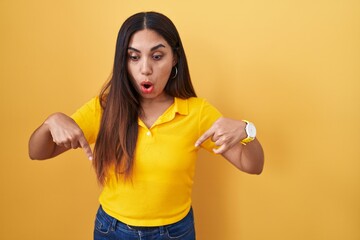 Young arab woman standing over yellow background pointing down with fingers showing advertisement,...