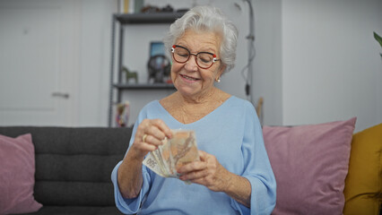 A cheerful senior woman counts czech koruna currency indoors, sitting in her cozy living room