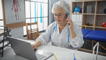 A senior woman healthcare professional consults a patient over the phone while working on a laptop...