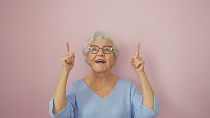 Joyful senior woman with glasses pointing upwards against a pink background.
