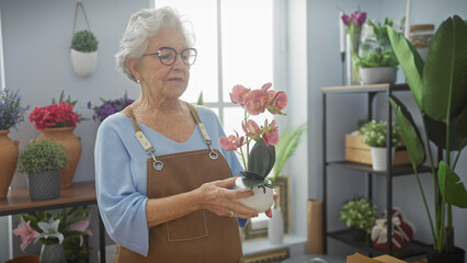 A senior woman with glasses holds an orchid in a modern flower shop with various plants.