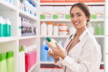 Young beautiful hispanic woman customer smiling confident holding shampoo bottles at pharmacy