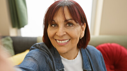 A cheerful middle-aged hispanic woman takes a selfie in a cozy, well-lit living room with a modern...