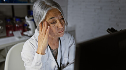 Middle age grey-haired lady scientist, focused face illuminated by the computerâ€™s glow,...