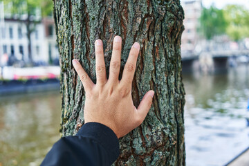 A person's hand touches the trunk of a tree by a city canal, suggesting a connection with nature in...