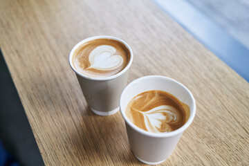 Two takeaway cappuccinos with heart latte art on a wooden table convey a cozy, coffee break atmosphere.