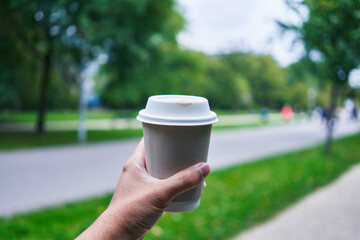 A man holds a takeaway coffee cup outdoors in a blurred park setting, symbolizing urban lifestyle...