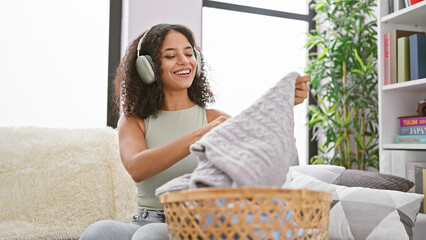Attractive young hispanic woman happily holding laundry basket full of clean clothing, enjoys music...
