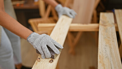 Hispanic woman carpenter at work, sanding wood plank with her hands at indoor carpentry workshop - obrazy, fototapety, plakaty