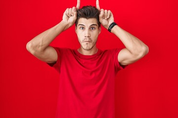 Fototapeta na wymiar Young hispanic man standing over red background doing funny gesture with finger over head as bull horns