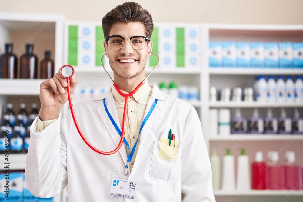 Sticker Young hispanic man working at pharmacy drugstore using stethoscope looking positive and happy standing and smiling with a confident smile showing teeth