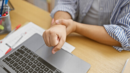 A man in an office winces in pain, clutching his wrist over a laptop, suggesting carpal tunnel...