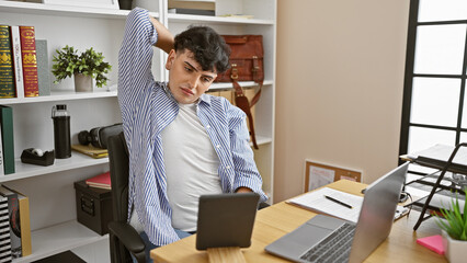 A young man stretches during a break in a modern office, displaying casual business attire and technology around.
