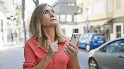 Mature caucasian woman holding smartphone looks pensive on sunny city street.